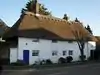 A wide, white-painted building with a prominent thatched roof and two chimneys. There are nine irregularly spaced windows; those at first-floor level are immediately below the roofline and are smaller. A blue door is set in the left-hand side, and a white gate is set next to it in a flint wall. A stumpy, possibly dead tree obscures the rightmost two windows.