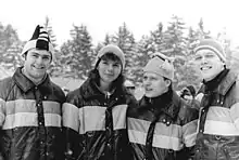 Three men and a woman are lined up side-by-side wearing winter caps and identical winter jackets. They are outdoors and snow is falling.