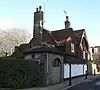 Almost side-on view of a long, two-storey house with two gable ends, large areas of tiles, a white ground floor and a flint section nearest the camera (with arched recess and brick buttress). Two tall chimneys rise from the roof. A low, narrow pavement with bollards runs alongside.