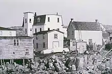 The photo shows a rocky foreground, then three clapboarded houses with assorted outbuildings. The Greenspond Courthouse, a large building with a central tower and mansard roof, is in the background.