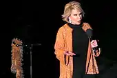 A woman with short blond hair wearing black except for a sparkly orange jacket with earrings. She is holding a microphone, and next to her is a microphone stand with an orange boa hanging from it.