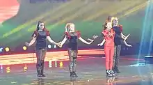 Manw in red standing on stage with four dancers in black standing diagonally behind her