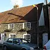 A small, two-storey flint cottage with a steep tiled roof, attached to another of the same height to the left. It has a white door and four irregularly spaced windows (edged in red brick), the upper two of which have window-boxes. A white gate is set into a flint wall attached to the right side of the cottage.