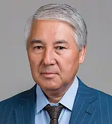 An older man of Central Asian complexion with grey hair, looking seriously into the camera with a grey background.