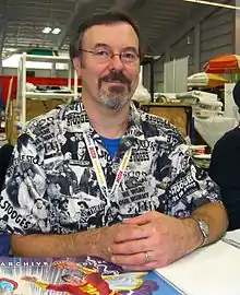 A middle-aged man with graying brown hair, a beard, glasses, and a Three Stooges shirt sitting at a table.