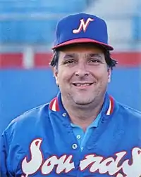 A man smiling wearing a blue satin jacket with "Sounds" across the front in white and red and a blue cap bearing a white "N"