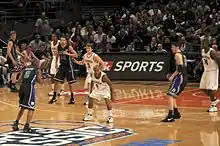 Two opposing basketball teams on a basketball court with a team in white defending the basket behind them. One team is in white uniforms with the word Michigan on the front and names on the back and the other is dark blue with the word Duke on the front and names on the back.