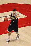 A basketball player in a dark blue uniform is dribbling the basketball and signalling with his index finger.