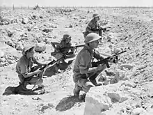 Soldiers crouch behind a low berm amidst a rocky desert area