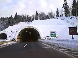 Tunnel portal, variable traffic signs indicating traffic flow direction and speed limit enforced are visible at the tunnel entrance and to the side of the road