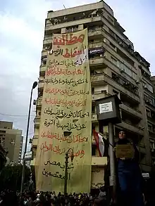 Large banner hanging from tall apartment building, with woman holding sign in foreground