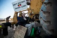 A woman sits with a chain around her waist, padlocked to heavy earthmoving equipment.