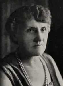 An older white woman with coiffed grey hair, wearing a dress or top with a low square neckline, and two strands of pearls