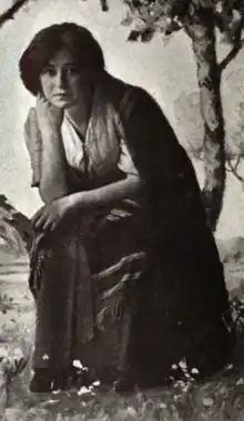 A white woman, seated and crouching, chin on hand, elbow on knee. She is wearing loose-fitting clothing, and appears to be in a stage set or studio background of trees and flowers.