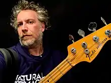 Alberto Rigoni close-up with greying hair and beard, looking at camera, wearing purple hoodie with lettering, holding up bass guitar with only head and neck showing