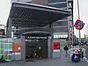 In the middle of building works a glass doors show banisters leading down beneath a sign reading "ALDGATE EAST STATION", this beneath a canopy supported on four girders.