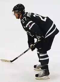 An ice hockey player leaning forward on his ice hockey stick and facing to the left of the camera. He is wearing a black helmet and uniform.