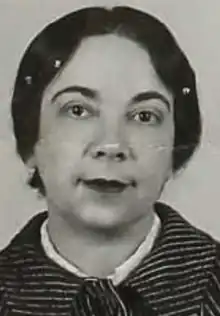 A middle-aged woman with fair skin and dark hair and eyes, wearing a white blouse unders a cloth coat with a striped or ribbed texture