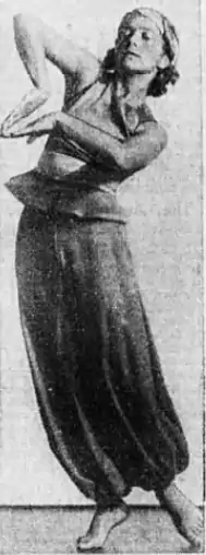 A young white woman standing barefoot in a costume with loose trousers, a sleeveless top with a peplum, and a headscarf. She is posing in a sway, with the backs of her hands facing each other, elbows poking outward.