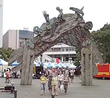 A wooden carved entrance arch in the middle of the photo with people walking through. Tents in the background from market day and pavers on the ground. The arch is traingular with two posts and is quite large. The carvings are a mixtere of shapes and some stick out of the top.