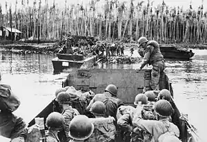 Black and white photo of a group of men wearing military uniform in an open-topped boat approaching a beach backed by palm trees. Two similar boats are ashore on the beach, with mean wearing military uniforms disembarking from one of them.