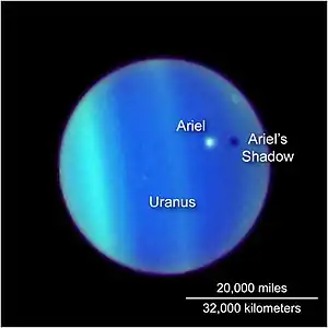 the planet Uranus is seen through the Hubble telescope, its atmosphere defined by bands of electric blue and green. Ariel appears as a white dot floating above it, casting a dark shadow below