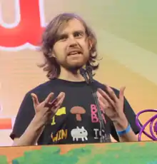 Arvi Teikari speaking behind a podium. He is wearing a black shirt with many icons from the game, including a flag, a mushroom, the "BABA", "IS", and "WIN" operators, and Baba itself.