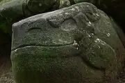 A smoothly finished boulder carved into the shape of a seated frog or toad, with the head raised to the right and sporting a prominent smile. The eyes, mouth, nostrils and legs are all carved in low relief. The sculpture is set against the stonework base of a structure, behind it at left.