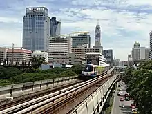 An elevated train, painted in blue, white and a red stripe and with advertisements, running above a road lined with many tall buildings and with many cars