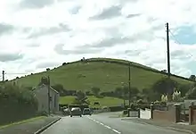 two-lane road bending downhill and to the right with a conical green hill beyond under a bright, cloudy sky