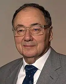 A clean-shaven older Caucasian man, his thin dark hair having receded to the middle of his head, looks at the camera through glasses with clear plastic rims; he is wearing a white shirt, grey patterned jacket and blue necktie.