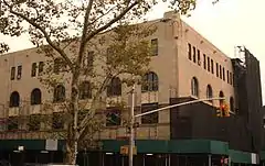 The corner of a rectangular six-story building is visible, partially obscured by a tree. The building is clad with slate colored stone, and at the top of the corner facing the viewer is a carving of a man holding stone tablets. The building sides have a row of rectangular windows, a row of arched windows above them, and a row of smaller rectangular windows above those. Much of the building, particularly the lower levels, is obstructed by construction hoarding, which also covers and protects the sidewalk adjacent.