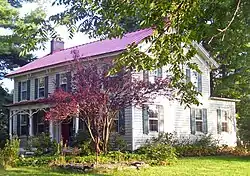 A white house with a red roof and green shutters partly obscured by some trees in front. It is lit from the right by late afternoon sunlight.