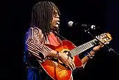 A man with dreadlocks wearing eyeglasses and a striped dress shirt; he is playing a guitar and standing behind a microphone stand