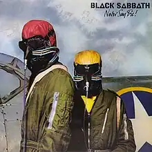 Two pilots stand in front of a fighter jet wearing forest green aviation jumpsuits. They each have odd-looking leather helmets, one colored tomato red, the other mustard yellow. Their faces are completely covered: Their eyes by dark goggles and their noses and mouths with black leather that has tubes attached for a breathing apparatus.