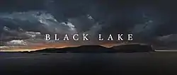 In the foreground, ice floats on the surface of a dark lake. In the background, snow caps the peaks of two mountains. The words Black Lake are superimposed in the centre.