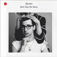 A picture of Jack Antonoff wearing a white T-shirt and a Star of David necklace. He rests his hand on his chin. An elderly woman places her hands on his hair, with her face out of frame.