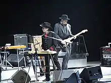 Dylan, dressed in a black western outfit with red highlights, stands onstage and plays the keyboards. He gazes to the left of the photo. Behind him is a guitar player, dressed in black.