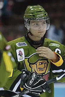 A frontal view of a white, teenage ice hockey player during a game. He is wearing a green, visored helmet and an olive green jersey with a logo consisting of an animated face of a soldier. He appears calm while looking to the right and is reaching for his face with his right hand.