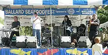 Four people on a stage; from left to right, they are playing a keyboard, a trumpet, a drum set, a guitar, and a saxophone. In the background are various signs, and on the stage are microphone stands and other audio equipment.