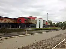 Broadhurst Park football ground on an overcast day, with an access road in the foreground. Visible are two white towers forming the front of the stadium, with a balcony to the left of the left tower. One of the unlit stadium floodlights is visible in the background.