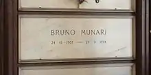 A white marble gravestone on the wall of a chapel, with only the name and dates of birth and death inscribed