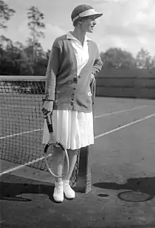 A woman looking away from the camera with a tennis racket in her right hand and a colored sweater on and all white clothing, which this picture is a black and white