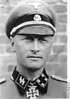 A man wearing a military uniform, peaked cap and a neck order in the shape of a cross. His cap has an emblem in shape of a human skull and crossed bones.