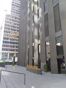 Entrances to the CBS Building, seen from Sixth Avenue looking north. Stairs lead up to the sidewalk to the west, and the entrances are to the east. There is a sculpted sign reading "Charles Schwab" in front of the entrances.