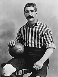 Head and upper torso of a young dark-haired man with thick eyebrows and a large moustache. He is looking straight ahead, and is wearing a tasselled cap and open-necked striped sports shirt.