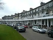 Row houses with continuous portico and stone walls on a curving street, with lines of cars parked in front