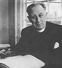 Canon Purvis, wearing ecclesiastical dress is sitting at a table facing the camera