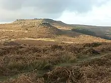 Photograph showing a moorland view. The moor is covered in heather of varying shades of brown. Stones are scattered across the moor. In the middle distance there is a rock outcrop atop a small hill. Behind it is a larger hill with a flat top.