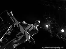 Diagonally tilted black-and-white photo of white male wearing a suit and hat, standing in front of a keyboard onstage, playing a violin.